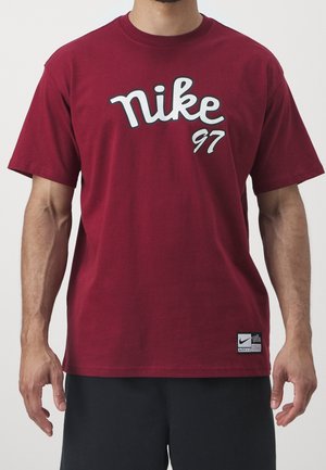 Mężczyzna ubrany w bordową koszulkę Nike z białym napisem „nike 97” i małą łatką z logo Nike przy dolnym brzegu, w zestawieniu z czarnymi spodniami.