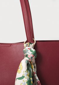 Anse de sac à main en cuir marron avec un anneau doré tenant une écharpe crème ornée de feuilles vertes et de fleurs jaunes, roses et rouges.