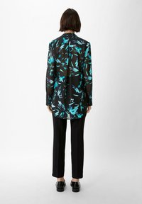 Blouse noire et transparente avec un motif abstrait bleu et turquoise ; manches longues, cou haut et un ourlet arrière légèrement plus long. Portée avec un pantalon noir.