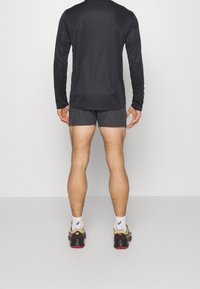 Schwarzes Langarm-Sportshirt kombiniert mit dunkelgrauen Shorts. Das Modell trägt schwarze und gelbe Laufschuhe mit weißen Socken. Glatte Textur.