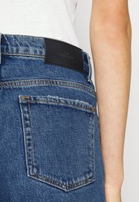 Jeans de denim azul con una cintura alta, que presentan un parche de cuero negro en la parte posterior y un bolsillo trasero con costuras visibles.