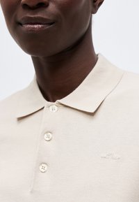 Κοντινό πλάνο ενός ατόμου που φοράει μπεζ πόλο μπλουζάκι με τρία κουμπιά και μικρό κεντημένο λογότυπο στο στήθος, με φόντο λευκό.