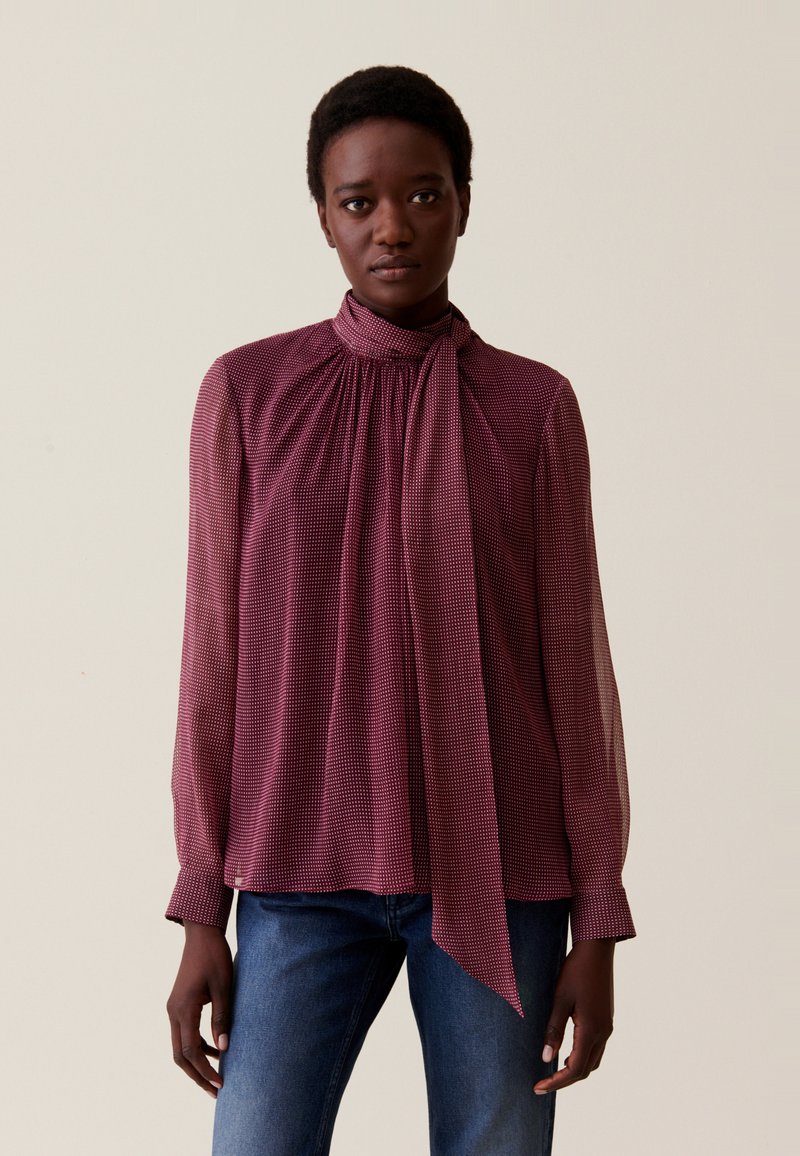 Blusa roja con estampado, de cuello alto y lazo, mangas largas y un ajuste relajado. Combinada con jeans azules. Tela suave con un brillo sutil.