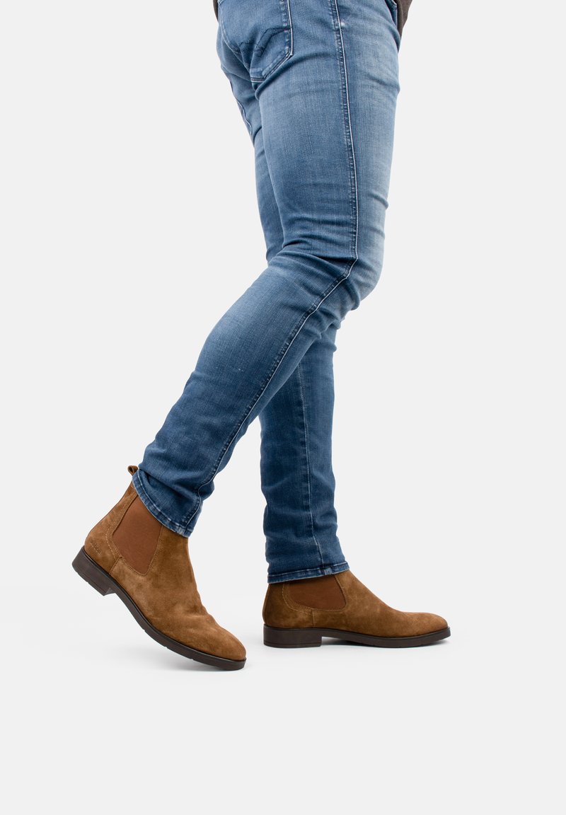 Botas Chelsea de ante marrón con paneles elásticos en los laterales y suelas de goma, combinadas con unos jeans de denim azul ajustados. Las botas tienen una punta redondeada.