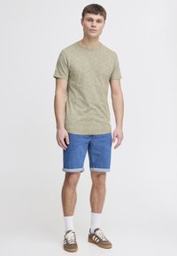 Olivengrønt kortærmet T-shirt med et tekstureret mønster, parret med blå denimshorts. Modellen har hvide sokker og brune sneakers på.