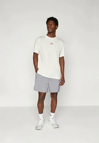 Άνδρας που στέκεται με τα χέρια στις τσέπες, φορώντας ένα λευκό μπλουζάκι Adidas, γκρίζες βερμούδες, λευκές κάλτσες και λευκά αθλητικά παπούτσια, μπροστά σε απλό φόντο.