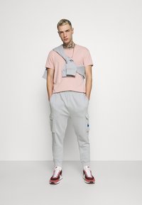 Svetloružové tričko s okrúhlym výstrihom, šedé tepláky s cargo vreckami a modrým logom, šedá mikina previazaná cez plecia, červené tenisky.