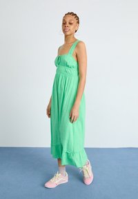 Robe midi en coton vert avec ourlet à volants et bretelles ajustables, associée à des baskets roses. Texture lisse, coupe décontractée, et détails de boutons à l'avant.