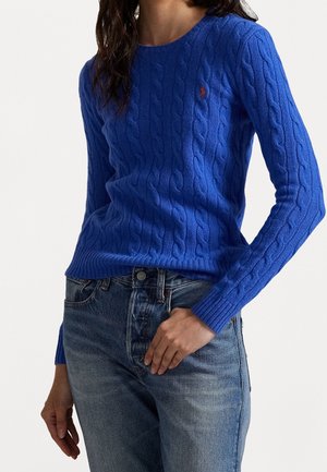 Mujer con un suéter ajustado azul de punto cable con mangas largas y jeans de tiro alto de color azul claro, con la mano apoyada cerca del bolsillo.