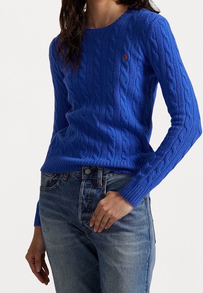 Femme portant un pull bleu moulant en maille torsadée avec manches longues et un jean taille haute bleu clair, la main reposant près de la poche.