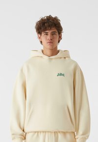 Cremefarvet hættebluse lavet af blødt stof, med en frontlomme og et grønt logo på brystet. Den har en hætte med snore.
