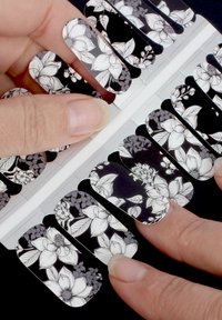 Wraps d'ongles floraux avec un fond noir et des motifs floraux blancs, en forme courbée, finition brillante et designs variés sur chaque wrap.