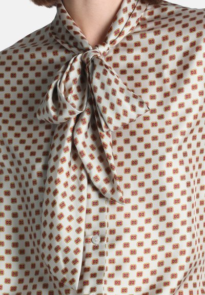 Blouse crème avec un motif géométrique rouge et jaune et un grand nœud noué au cou.