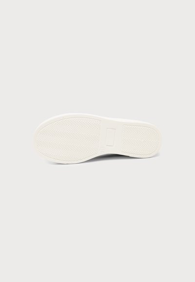 Semelle de chaussure blanche avec motif en arêtes de poisson, détail rectangulaire près de la voûte plantaire, photographiée sur un fond gris clair.