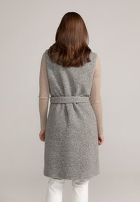 Langer grauer ärmelloser Mantel aus einem weichen Wollmix, mit tailliertem Bund, Rückenschlitz und glatter Textur. Getragen über einem leichten Strickoberteil.