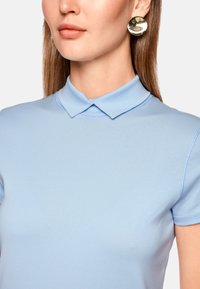 Light blue polo shirt with a soft texture, features a classic collar and short sleeves, paired with shiny gold circular earrings.