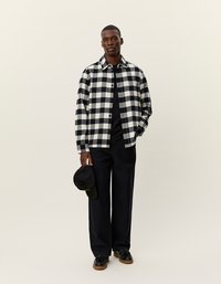 Fekete-fehér kockás flanel ing, alatta fekete póló, sötét nadrág, kezében fekete sapka, és sötét cipő. Minimalista háttér.