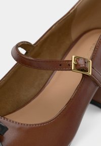 Chaussure Mary Jane en cuir marron avec une bride à boucle dorée et un talon bloc, montrant un gros plan du côté et de la doublure intérieure.