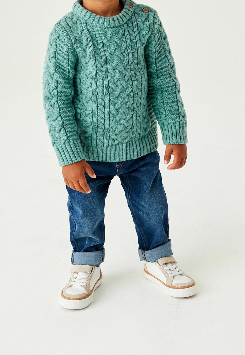 Enfant portant un pull torsadé bleu sarcelle, un jean bleu foncé retroussé et des baskets blanches sur fond blanc.