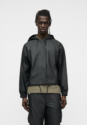 Άνδρας που φορά μαύρο μπουφάν με κουκούλα και μαύρο παντελόνι cargo πάνω από μια χακί πράσινη μπλούζα, στέκεται μπροστά σε ένα απλό ανοιχτό γκρι φόντο.