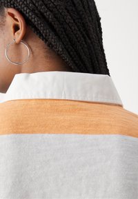 Kurzärmliges Oberteil mit einem orange-grauen Farbblock-Design. Das Material wirkt weich, mit einem knallweißen Kragen; beinhaltet einen großen silbernen Creolen-Ohrring.