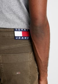 Pantalones verde oliva con un acabado texturizado y un bolsillo trasero. Presentan una etiqueta azul con "TOMMY JEANS" y un panel con acento rojo/blanco.