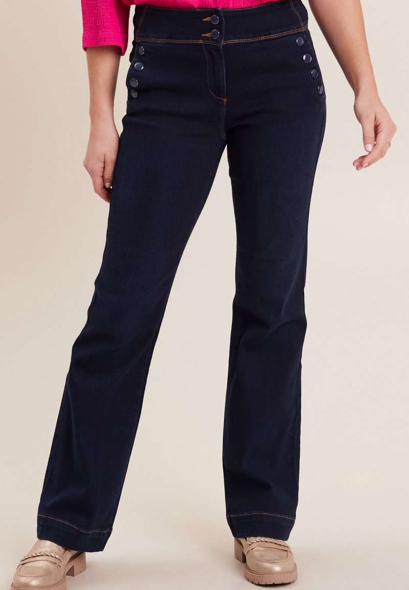 Jeans a campana blu scuro con cinque bottoni sulla parte anteriore, cuciture arancioni a contrasto e una texture di denim liscia. Abbinati a scarpe chiare.