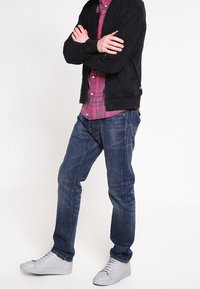 Ciemnoniebieskie dżinsy o prostym kroju noszone z czarną kurtką bomberką na różowej koszuli w kratę, w połączeniu z białymi sneakersami.