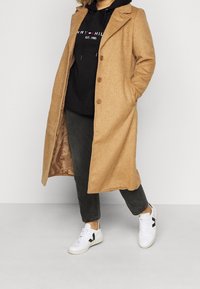 Lichtbruine wolblend jas met een geknipte kraag, knoopsluiting en zijzakken, gelaagd over een zwarte hoodie en grijze broek, gecombineerd met witte sneakers.