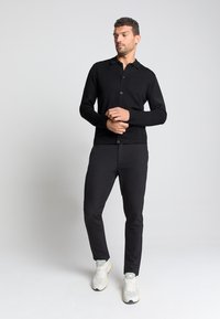 Zwarte poloshirt met lange mouwen, kraag en knoopsluiting, gecombineerd met zwarte broek en witte sportieve schoenen. Glad textuur en aansluitend ontwerp.