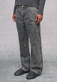 Jeans de mezclilla gris que presentan un corte relajado con un patrón de desgaste prominente. Combinados con elegantes zapatos gris de tipo slip-on.
