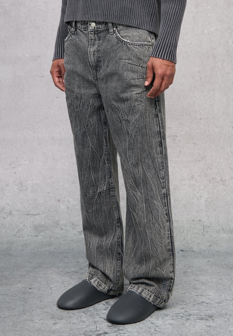 Jeans de mezclilla gris que presentan un corte relajado con un patrón de desgaste prominente. Combinados con elegantes zapatos gris de tipo slip-on.