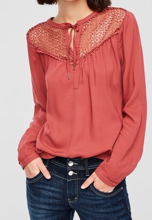 Kobieta ubrana w rdzawobrązową bluzkę z długim rękawem, z koronkowym karczkiem i wiązaniem przy dekolcie, z rękami w kieszeniach ciemnoniebieskich dżinsów z podwójnym zapięciem na guziki.