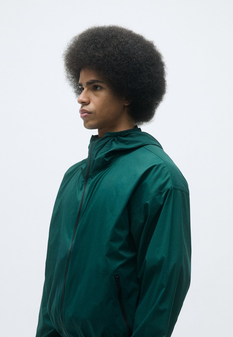 Grüne, leichte Jacke mit hohem Kragen und Reißverschluss, mit glatter Textur und Seitentaschen. Das Modell hat eine Afro-Frisur.