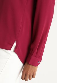 Bordeaux blouse van een gestructureerde stof. Beschikt over een lange mouw, knoop manchetten en zijinkepingen voor extra detail. Gecombineerd met witte pantalons.