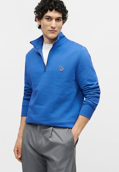 Άνδρας με μαύρα σγουρά μαλλιά, φοράει μπλε φούτερ με φερμουάρ μέχρι το στήθος και μικρό λογότυπο ζώου και γκρι παντελόνι, με το ένα χέρι στην τσέπη.