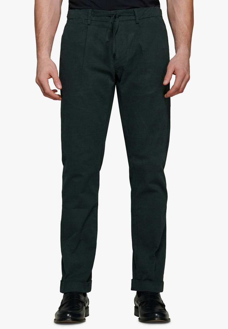 Dunkelgrüne Hosen mit texturiertem Muster, schmal zulaufendem Design, Kordelzugbund und aufgerollten Säumen, kombiniert mit schwarzen Schuhen.