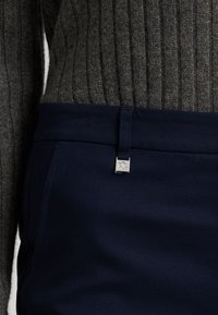 Pantaloni navy in un tessuto liscio presentano un'etichetta in metallo argento con "RL," abbinati a un maglione grigio scuro a coste e texture.