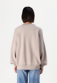 Osoba s krátkými tmavými vlasy má na sobě volný béžový svetr a modré džíny, stojí zády k jednobarevnému bílému pozadí.