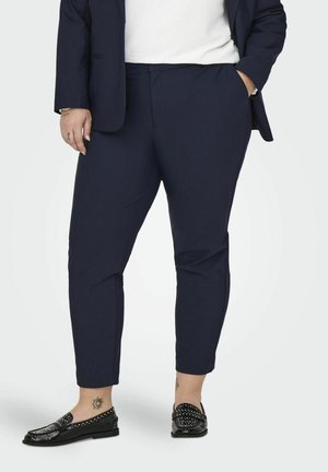Marineblaue maßgeschneiderte Hose mit geradem Schnitt, seitlichen Taschen und glatter Textur, kombiniert mit schwarzen Mokassins mit Nietenakzenten.