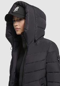 Doudoune noire avec une grande capuche, fermeture éclair sur le devant et tissu texturé. Présente un logo sur l'épaule et une casquette noire assortie.