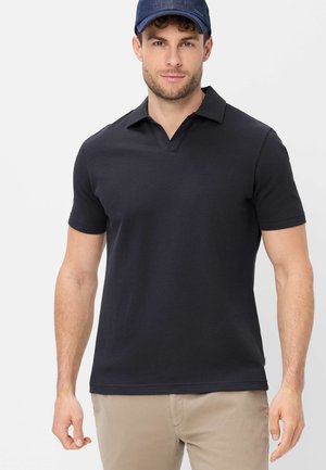 Uomo che indossa una polo a maniche corte di colore blu navy scuro con texture, pantaloni beige e un berretto blu navy, in piedi davanti a uno sfondo chiaro e uniforme.