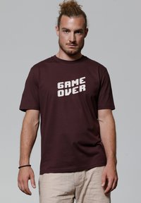 Bordeauxrood T-shirt van katoen, met een witte pixel-achtige "GAME OVER" graphic op de voorkant. Korte mouwen en een standaard ronde hals.