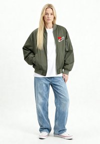 Olijfgroene bomberjack met een rode hart- en veiligheidsspeld-borduring, ritssluiting aan de voorkant, geribbelde manchetten, gecombineerd met losse blauwe jeans en witte sneakers.