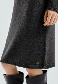 Robe en maille grise à coupe droite, avec des poignets côtelés et une petite étiquette dorée en bas. La texture semble douce et lisse.