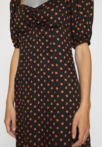 Robe noire avec un petit motif floral orange, bustier froncé et courtes manches bouffantes portée par une personne avec les mains relâchées sur les côtés.