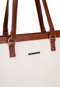 Bolso tote texturizado blanco con acentos de cuero marrón, herrajes dorados y un logotipo de marca prominente en la parte frontal. Cuenta con dos asas marrones.