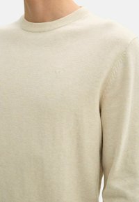 Maglione a maglia beige chiaro con collo a giro, caratterizzato da una superficie testurizzata e un sottile logo sul petto. Materiale morbido e leggero.