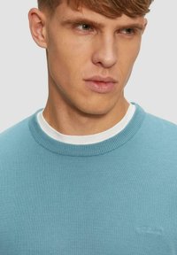 Lichtblauwe gebreide trui met een ronde hals en ribbelstructuur. Kenmerkt zich door een witte kraag eronder en een klein logo op de borst.
