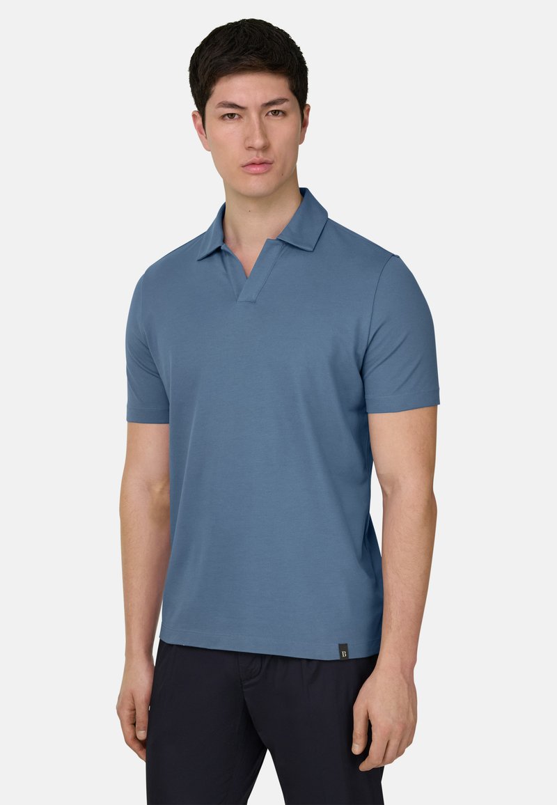 Uomo con polo a maniche corte blu e pantaloni scuri, in piedi su uno sfondo bianco uniforme.